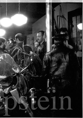 Image description: Men in leather jackets crowd into a small, lamp-lit shop where art hangs on the wall. Boots are on the shelves and in the display window. Outside the window stands a motorcyle. A leatherman wearing a bikers' cap is entering the shop; his keys hang on the left.